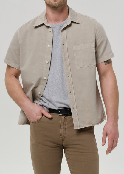 Person wearing a light beige short sleeve button-up shirt with brown straight leg pants on a grey background.