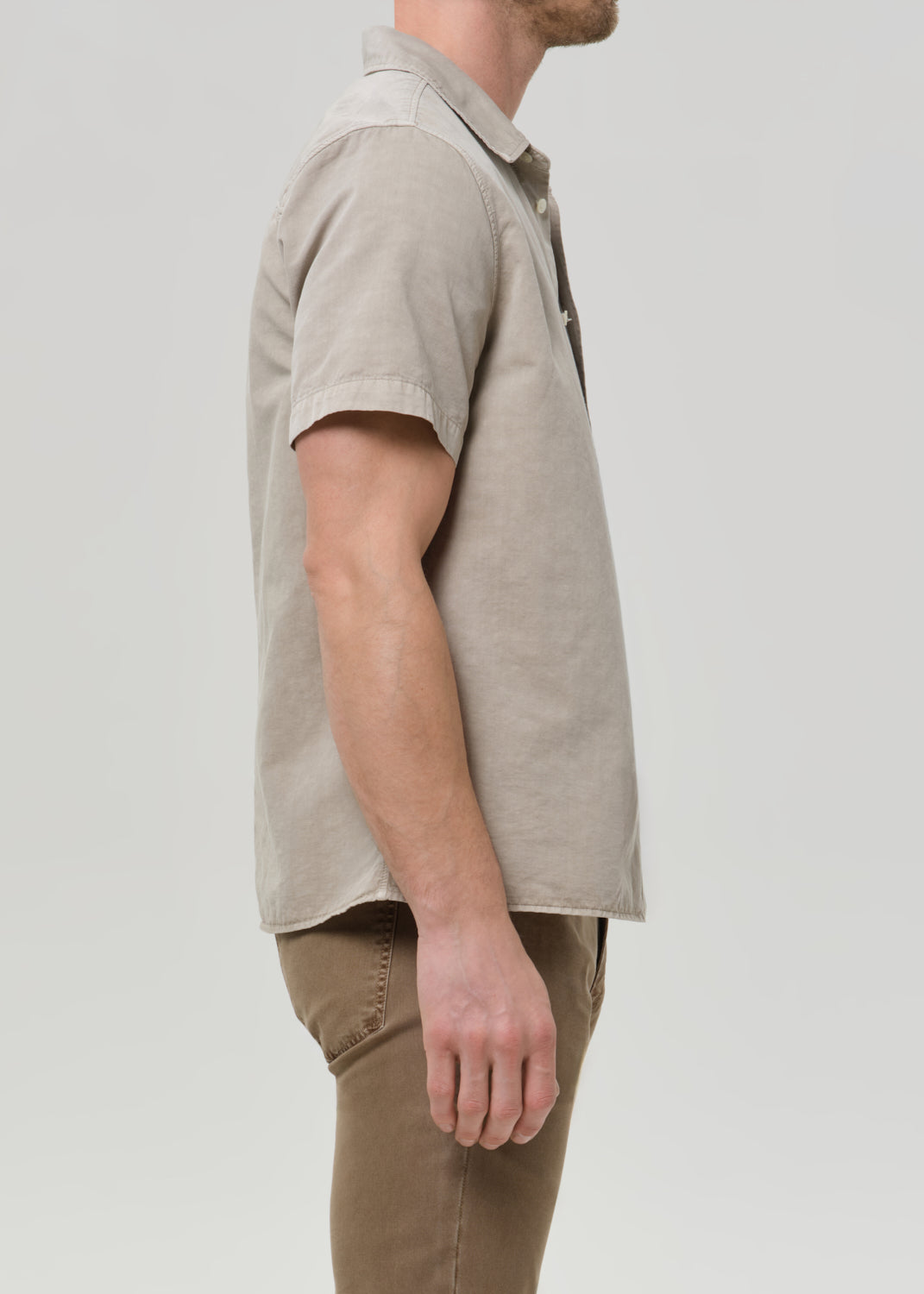 Person wearing a light beige short sleeve button-up shirt with brown straight leg pants viewed from the side on a grey background.