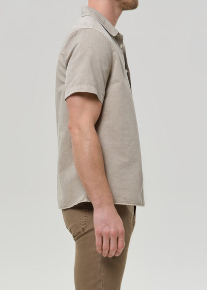 Person wearing a light beige short sleeve button-up shirt with brown straight leg pants viewed from the side on a grey background.