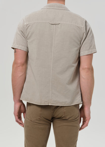 Person wearing a light beige short sleeve button-up shirt with brown straight leg pants viewed from the back on a grey background.