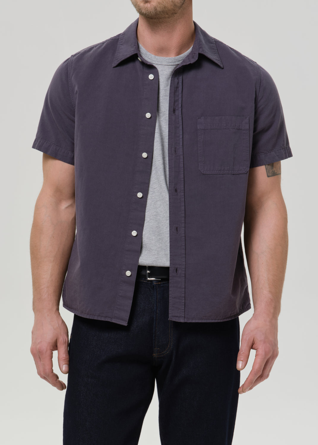 Front view of a man wearing a dark purple short-sleeve button-up shirt and dark jeans against a grey background.