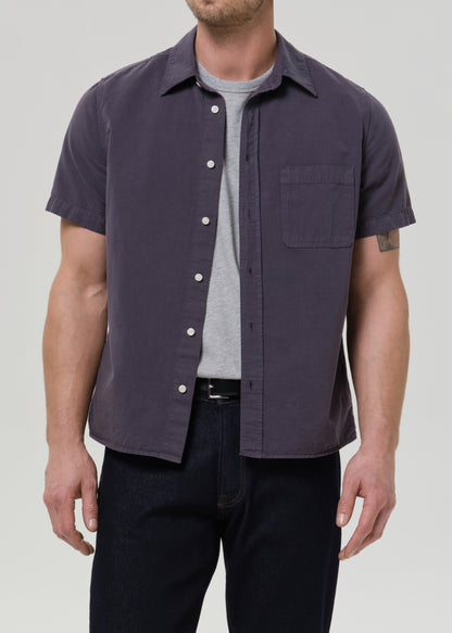 Front view of a man wearing a dark purple short-sleeve button-up shirt and dark jeans against a grey background.