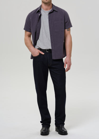 A man modeling a dark purple short-sleeve button-up shirt over a gray t-shirt, paired with dark navy pants and black shoes, against a grey background.