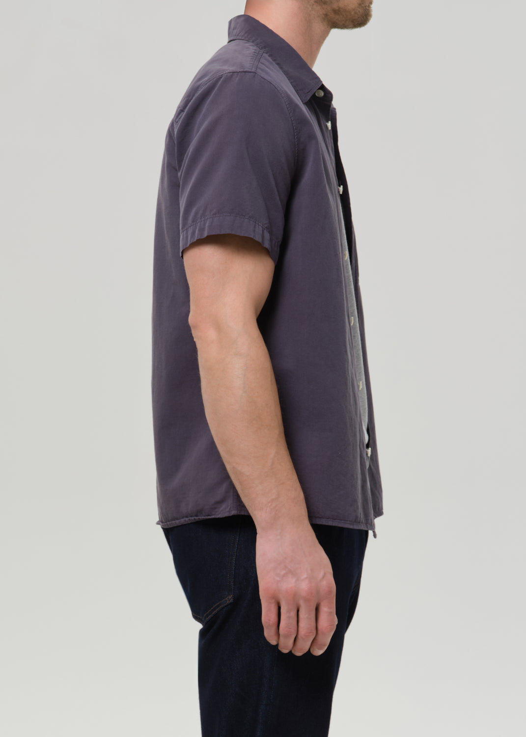 Side profile of a man wearing a short-sleeve button-up shirt in a muted dark purple-gray color, paired with dark jeans, against a grey background.