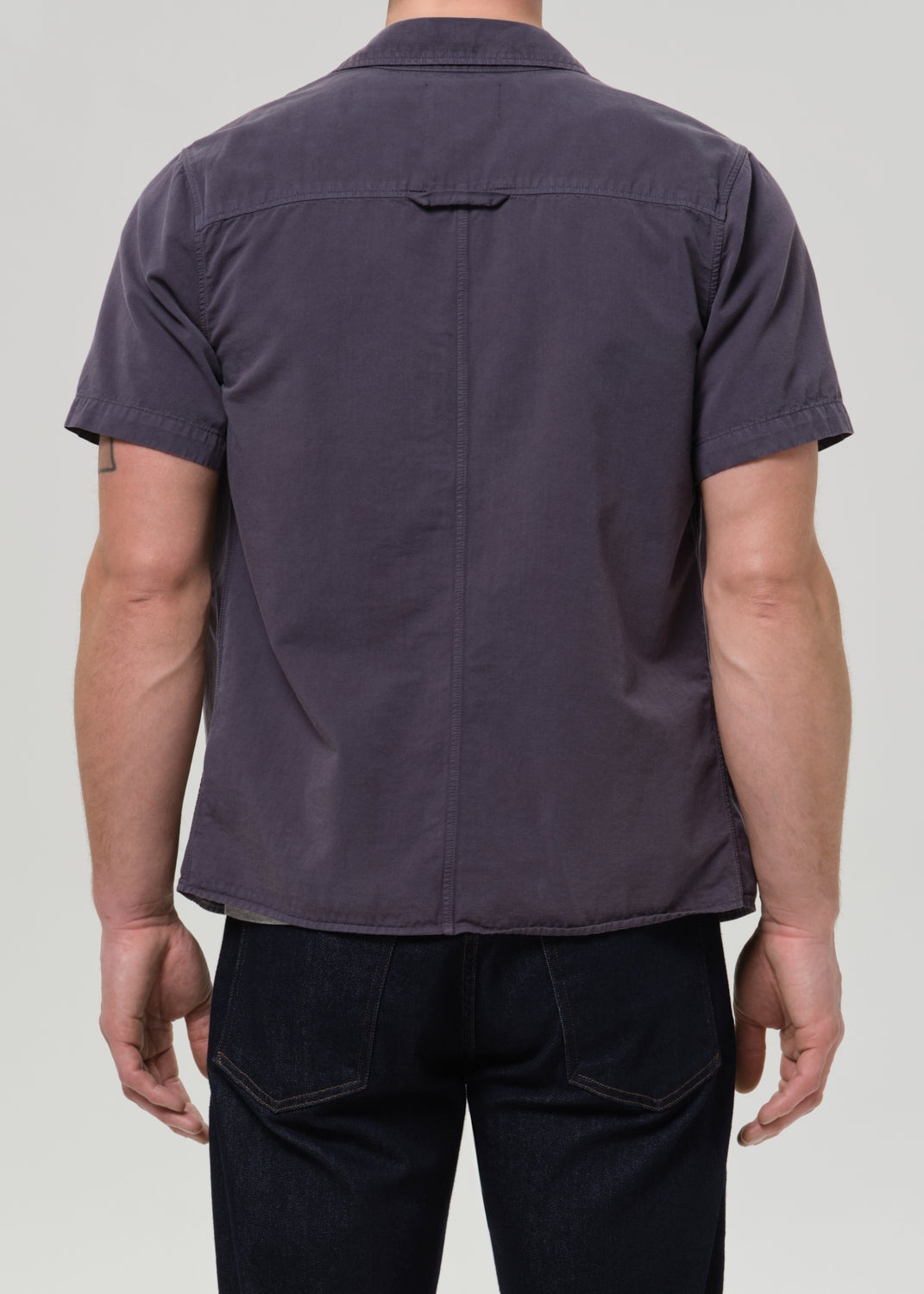 Rear view of a man wearing a dark purple short-sleeve button-up shirt and dark jeans against a grey background.