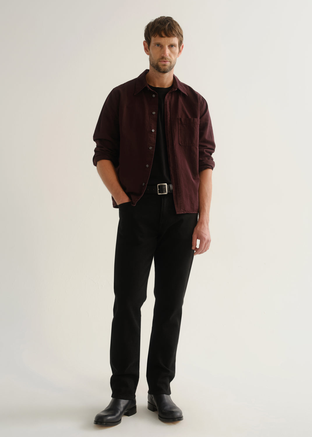 Person wearing a dark burgundy button-up shirt over a black shirt with black pants and black shoes, front full body view on a light grey background.