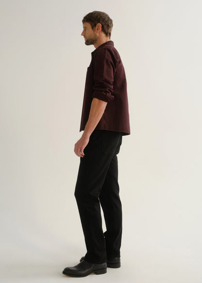 Person wearing a dark burgundy button-up shirt with black pants and black shoes, side full body view on a light grey background.