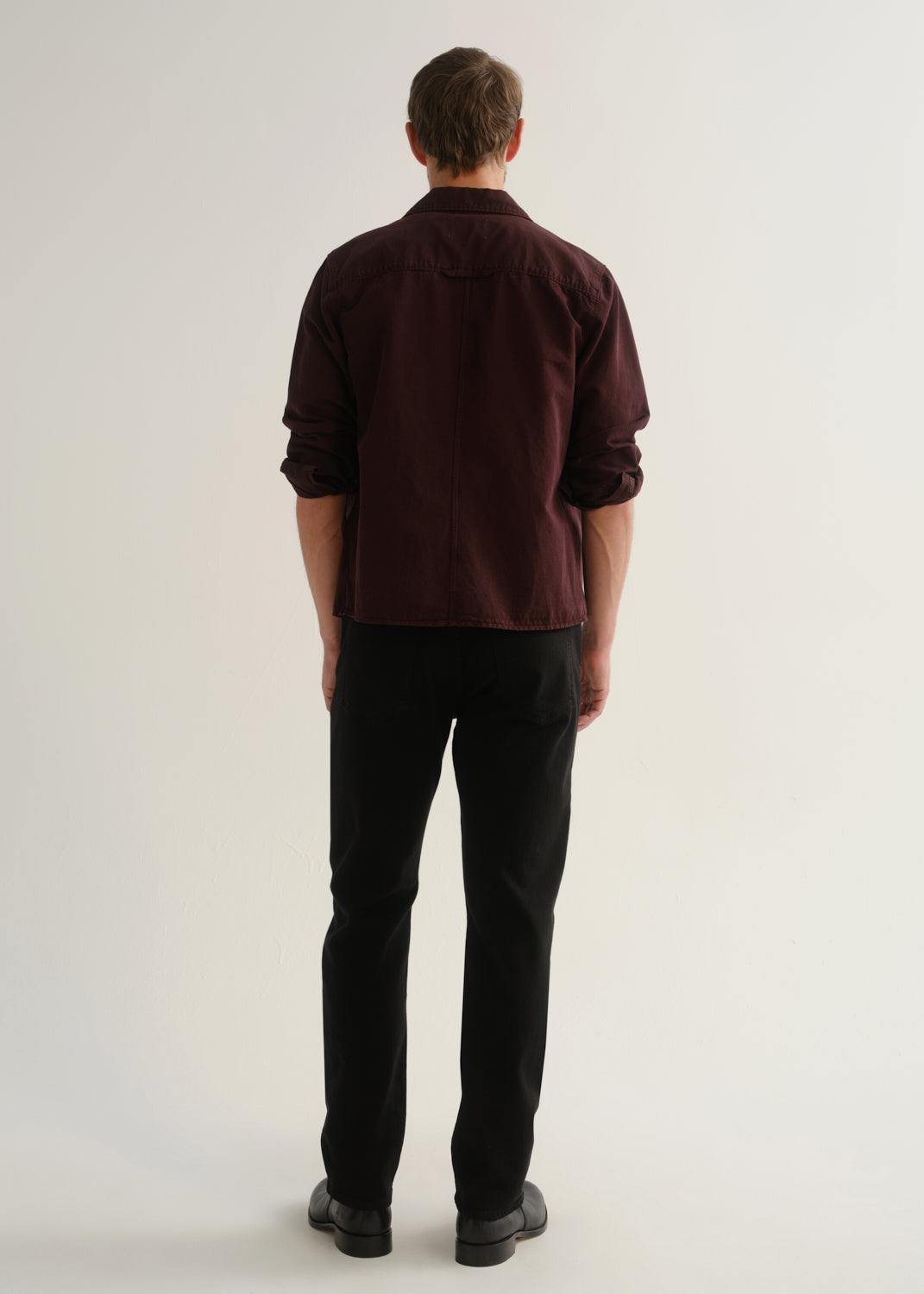 Person wearing a dark burgundy button-up shirt with black pants and black shoes, back full body view on a light grey background.