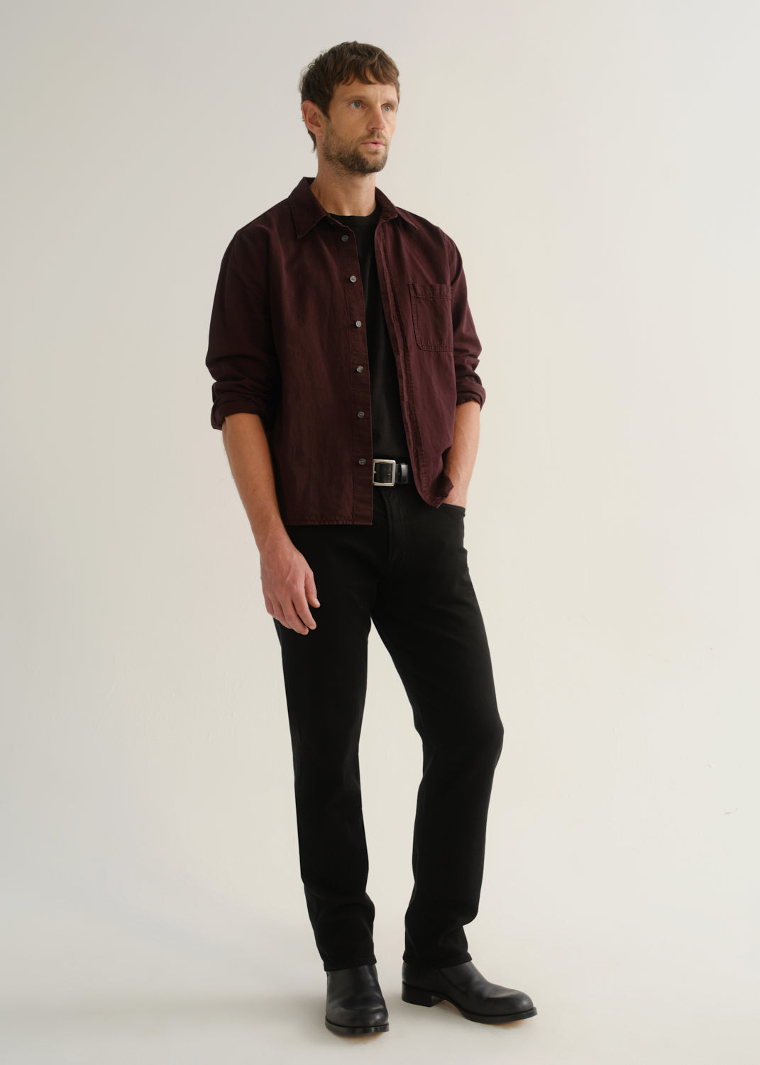 Person wearing a dark burgundy button-up shirt over a black shirt with black pants and black shoes, angled front full body view on a light grey background.
