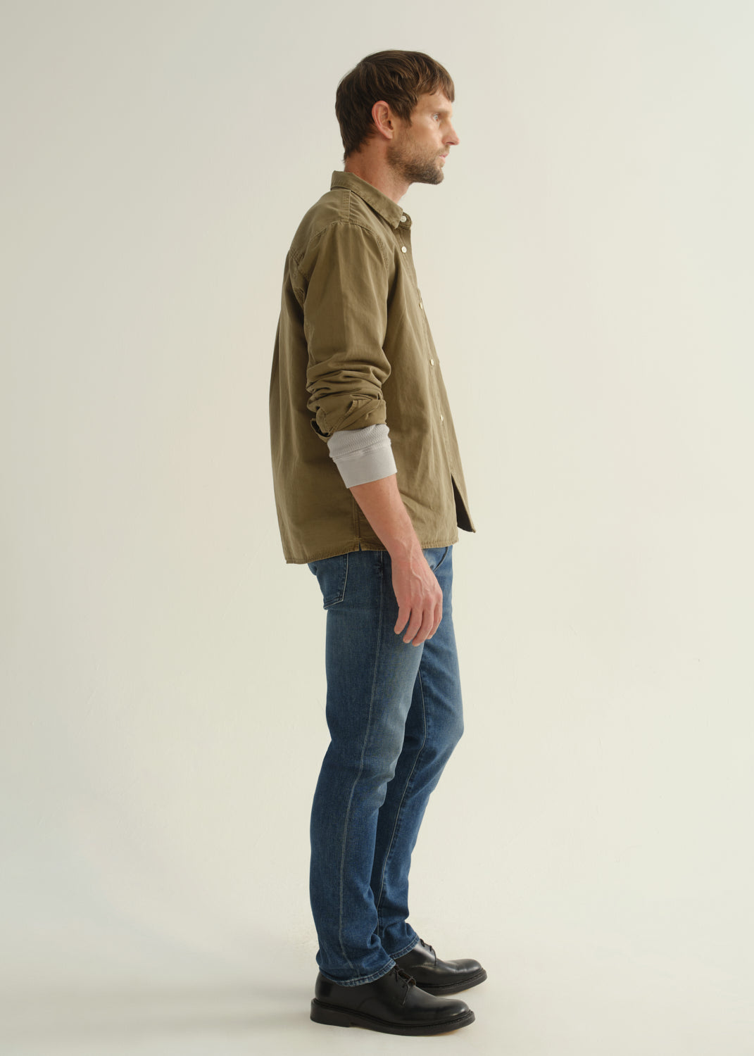 Person wearing an olive khaki long sleeve button-up shirt, blue jeans, and black boots, side full body view on a light grey background