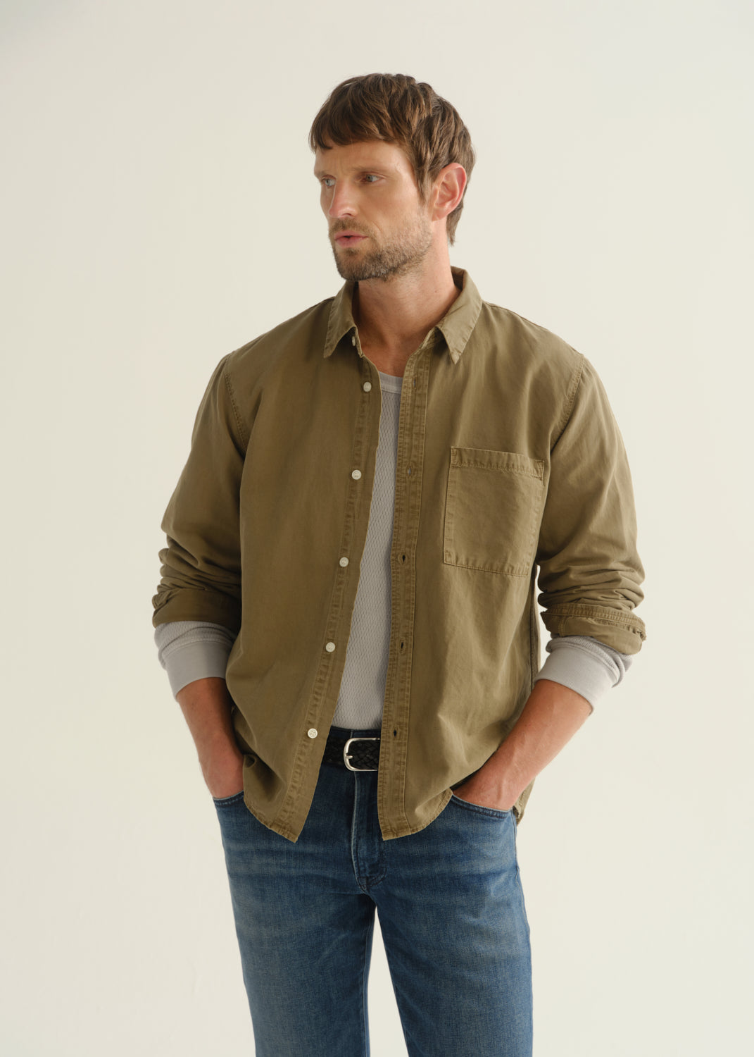 Person wearing an olive khaki long sleeve button-up shirt, a white t-shirt, and blue jeans, front cropped view on a light grey background.