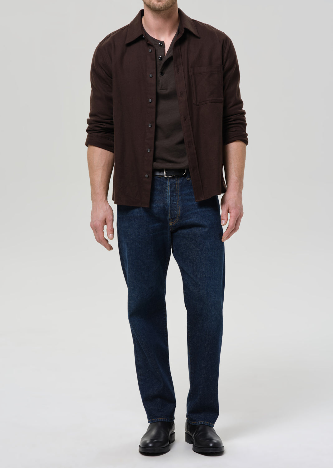 Man wearing a brown button-up shirt and blue jeans on a grey background