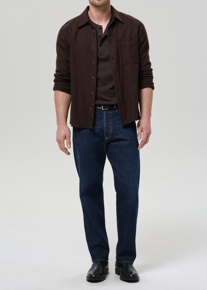 Man wearing a brown button-up shirt and blue jeans on a grey background