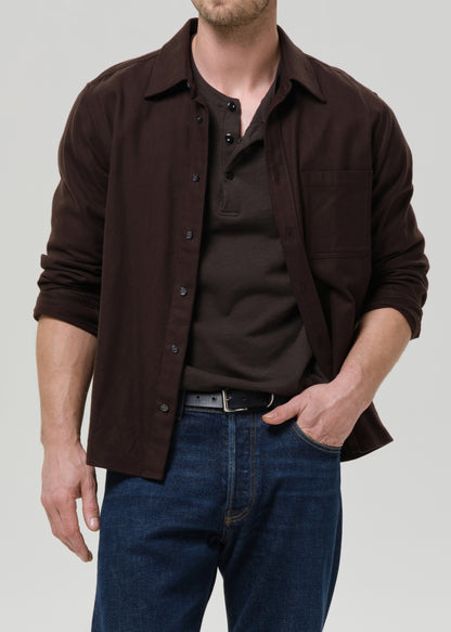 Man wearing a brown shirt over a black shirt with blue jeans on a grey background