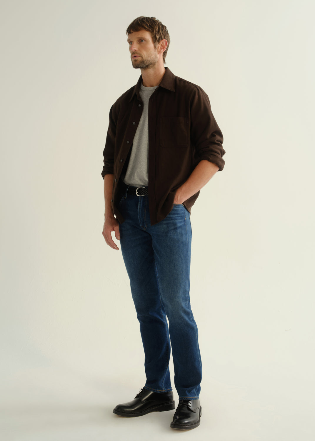 Person wearing a dark brown button-up shirt over a grey shirt with blue jeans and black shoes, angled front full body view on a light grey background.