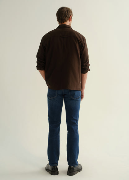 Person wearing a dark brown button-up shirt with blue jeans and black shoes, back full body view on a light grey background.