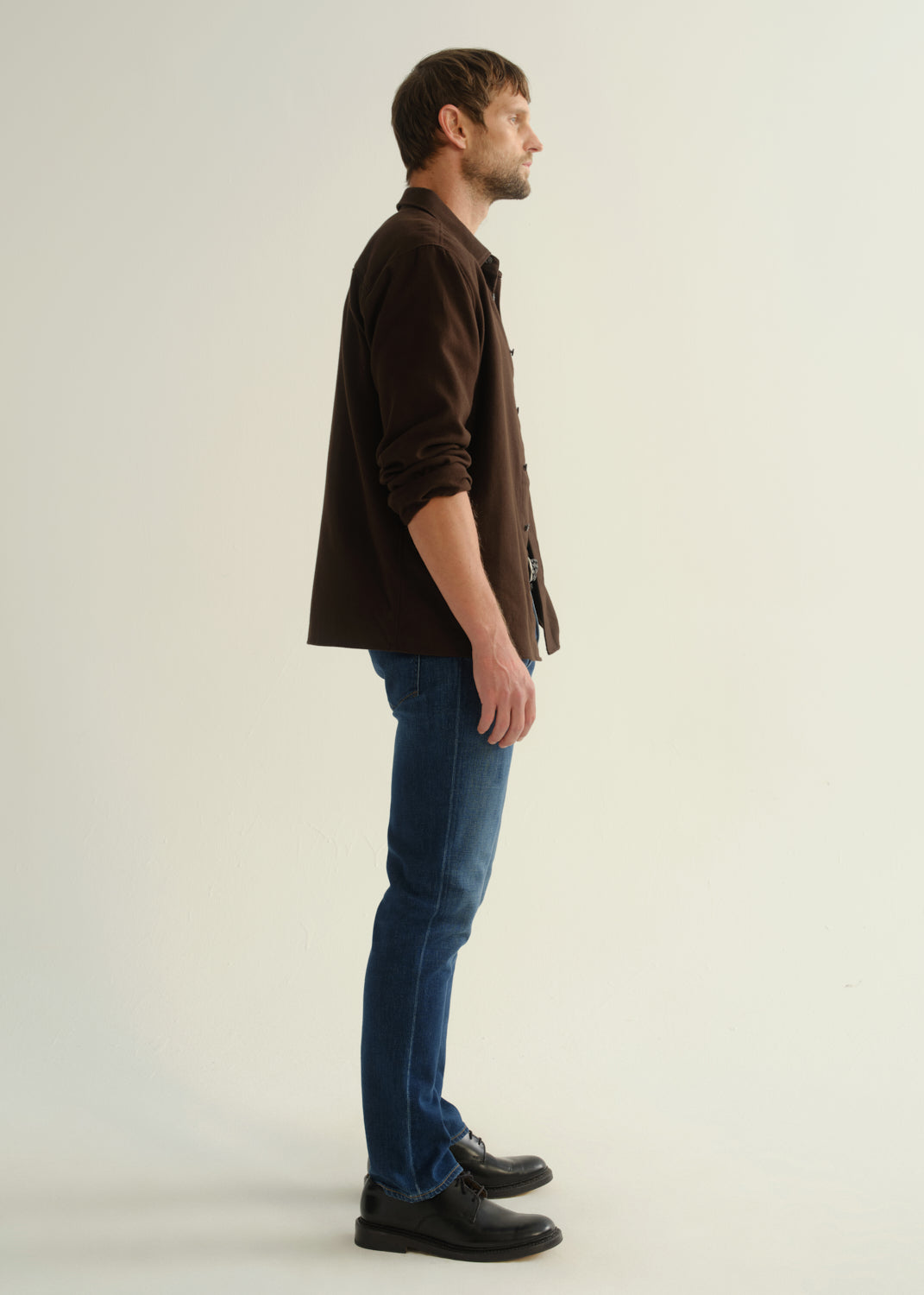 Person wearing a dark brown button-up shirt with blue jeans and black shoes, side full body view on a light grey background.