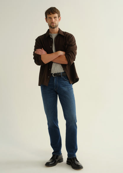 Person wearing a dark brown button-up shirt over a grey shirt with blue jeans and black shoes, front full body view on a light grey background.