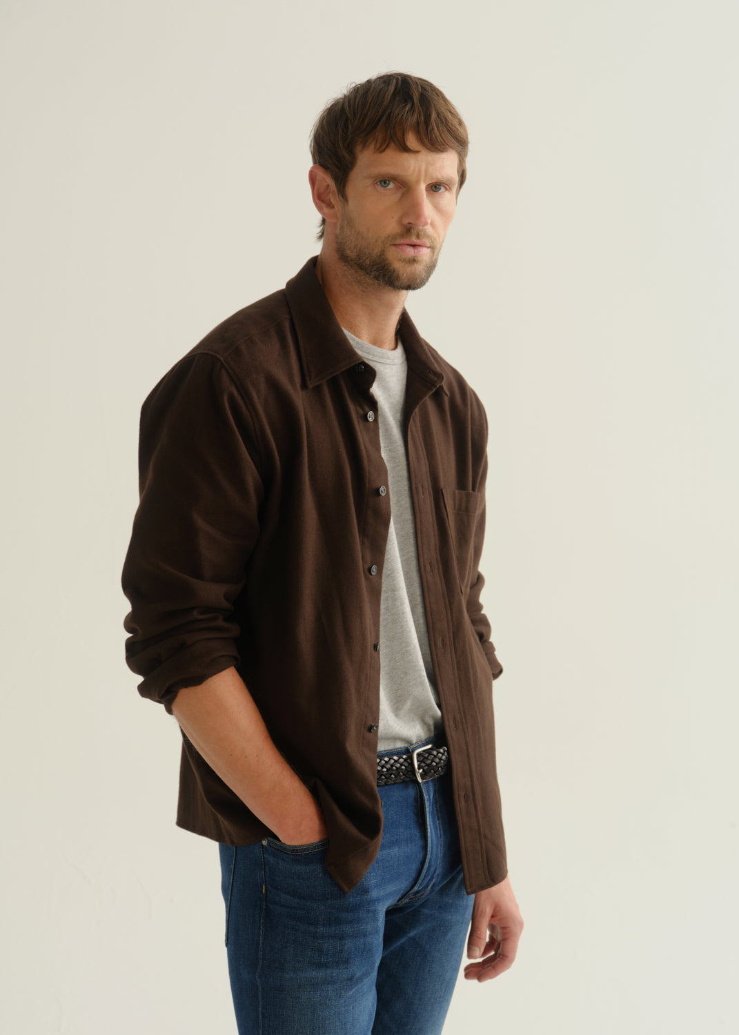 Person wearing a dark brown button-up shirt over a grey shirt with blue jeans, front cropped view on a light grey background.