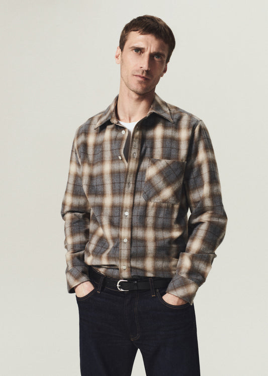 Man wearing a brown and grey plaid button-up shirt layered over a white crewneck top with dark indigo jeans on a grey background.