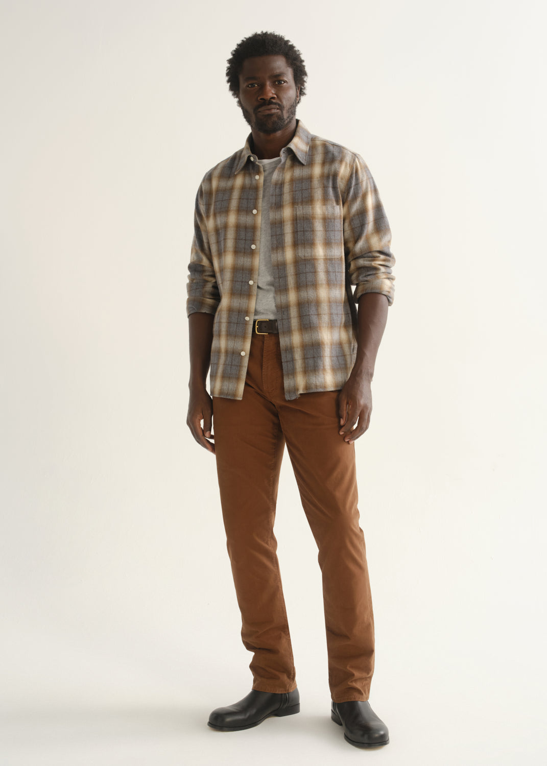 Person wearing a multicolor plaid button-up shirt over a grey t-shirt with brown pants and black shoes, front full body view on a light grey background.