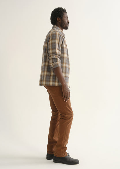 Person wearing a multicolor plaid button-up shirt with brown pants and black shoes, side full body view on a light grey background.
