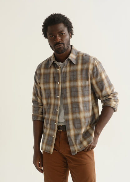 Person wearing a multicolor plaid button-up shirt over a grey t-shirt with brown pants, front cropped view on a light grey background.