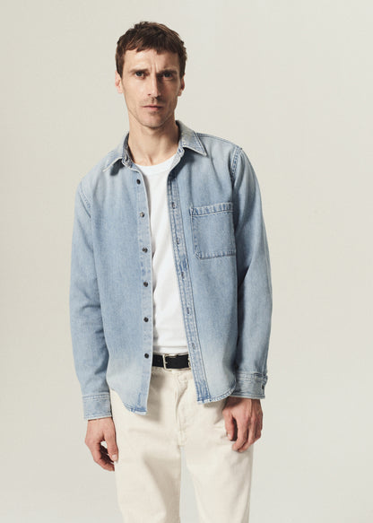 Man wearing a light blue denim button-up shirt layered over a white crewneck top with ecru pants on a grey background.