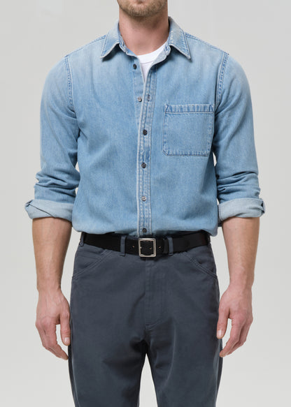 Man wearing a light blue denim shirt and dark pants on a grey background