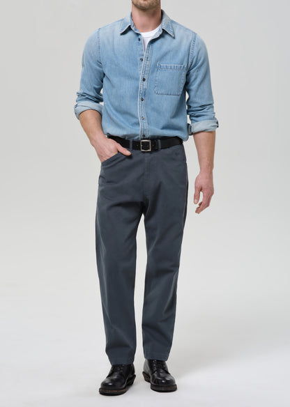Man wearing a light blue denim shirt and dark gray pants on a grey background