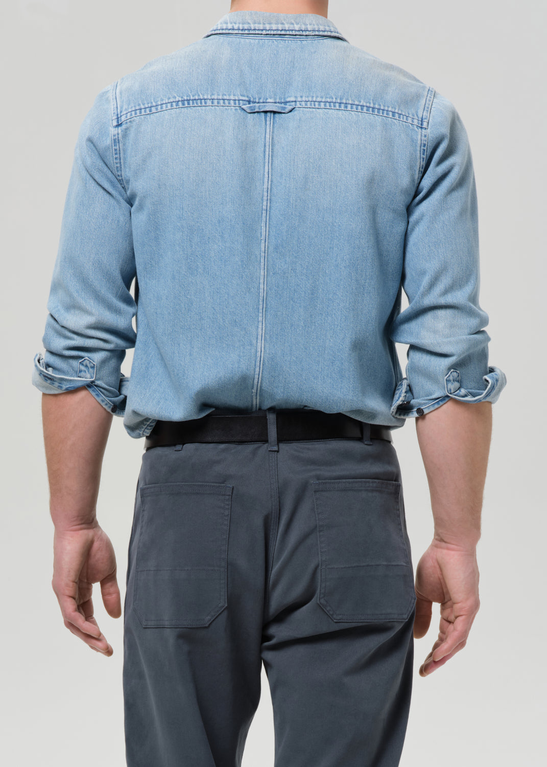 Person wearing a light blue denim shirt and dark grey pants on a grey background