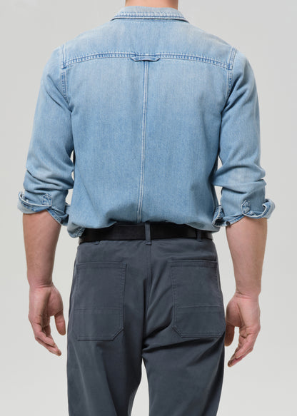 Person wearing a light blue denim shirt and dark grey pants on a grey background