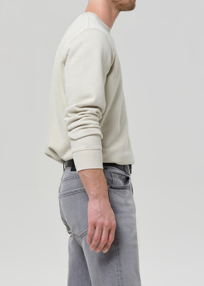 Person wearing a cream crewneck sweatshirt with light grey straight leg jeans viewed from the side on a grey background.