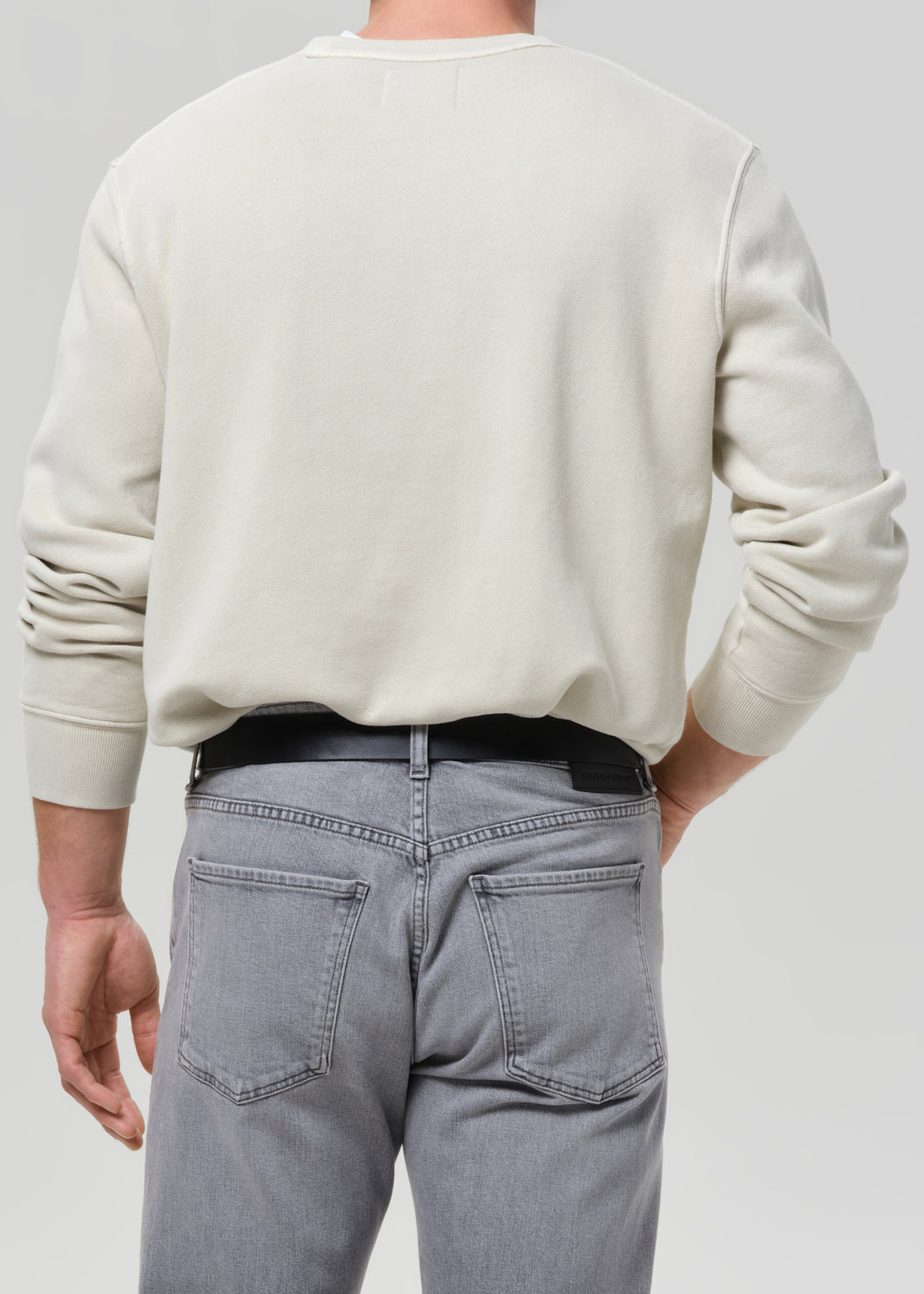 Person wearing a cream crewneck sweatshirt with light grey straight leg jeans viewed from the back on a grey background.