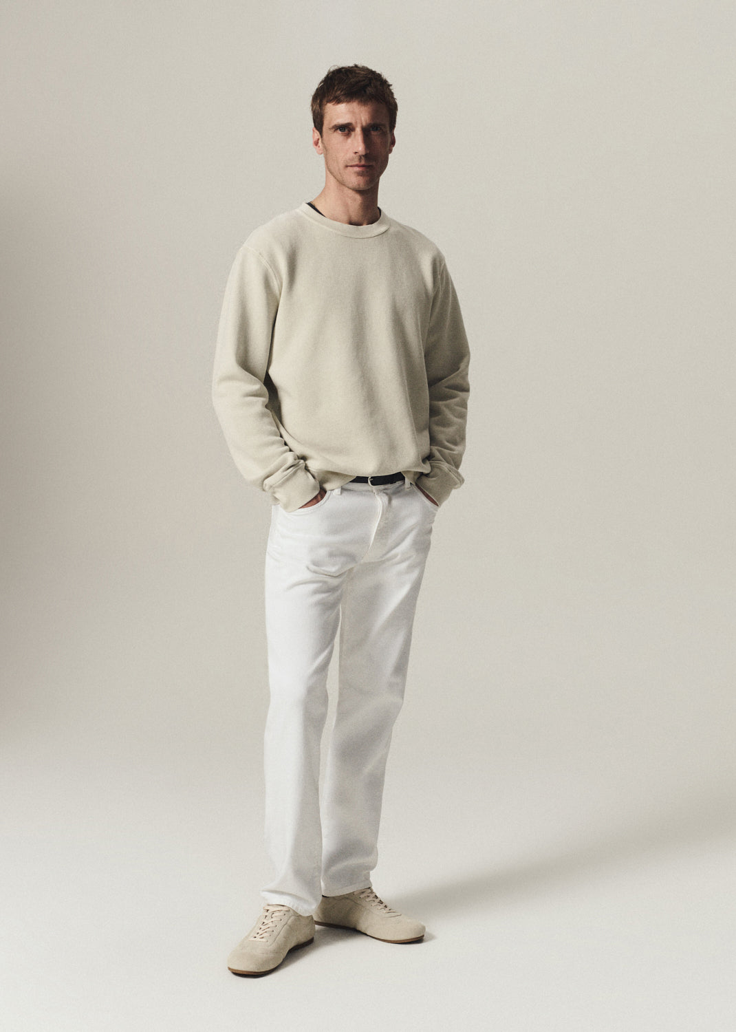 A man modeling a light beige crewneck sweatshirt, white trousers, and beige sneakers against a plain, warm off-white studio background.