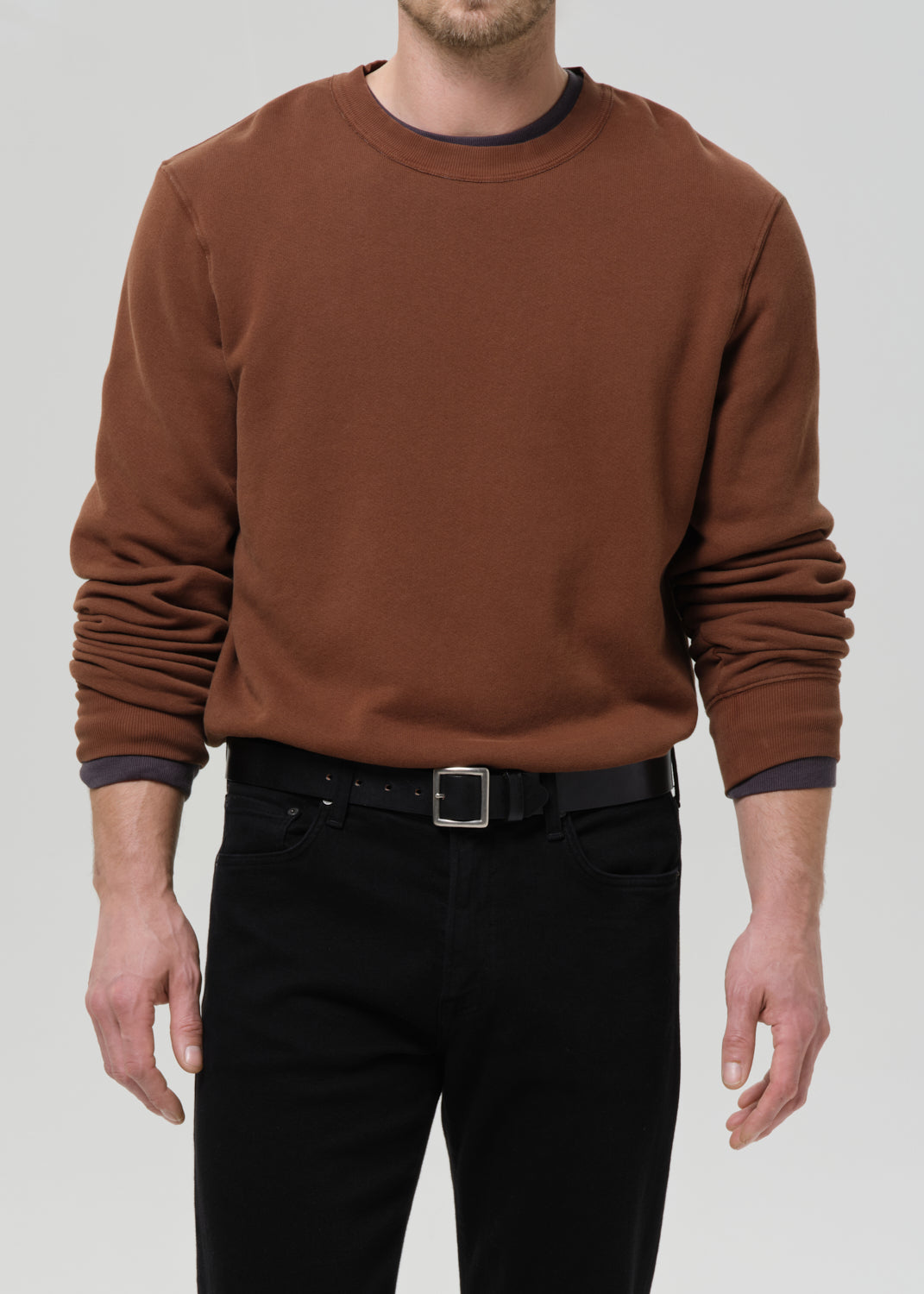Person wearing a brown crewneck sweatshirt with black straight leg jeans on a grey background.