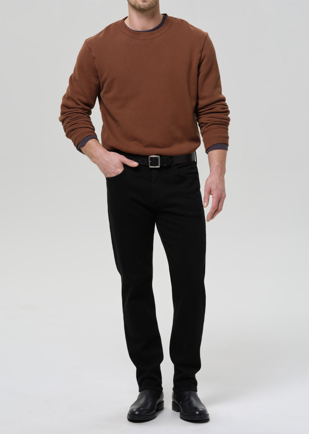 Person wearing a brown crewneck sweatshirt with black straight leg jeans on a grey background.