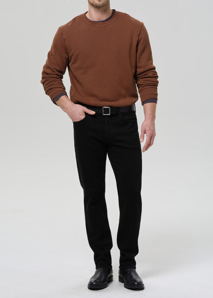 Person wearing a brown crewneck sweatshirt with black straight leg jeans on a grey background.