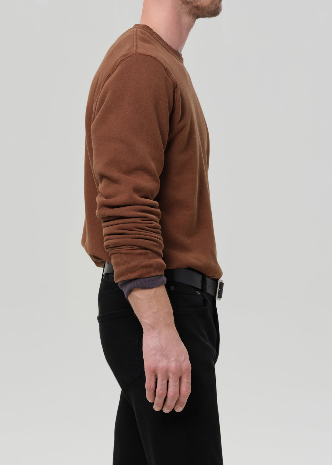 Person wearing a brown crewneck sweatshirt with black straight leg jeans viewed from the side on a grey background.