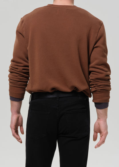 Person wearing a brown crewneck sweatshirt with black straight leg jeans viewed from the back on a grey background.