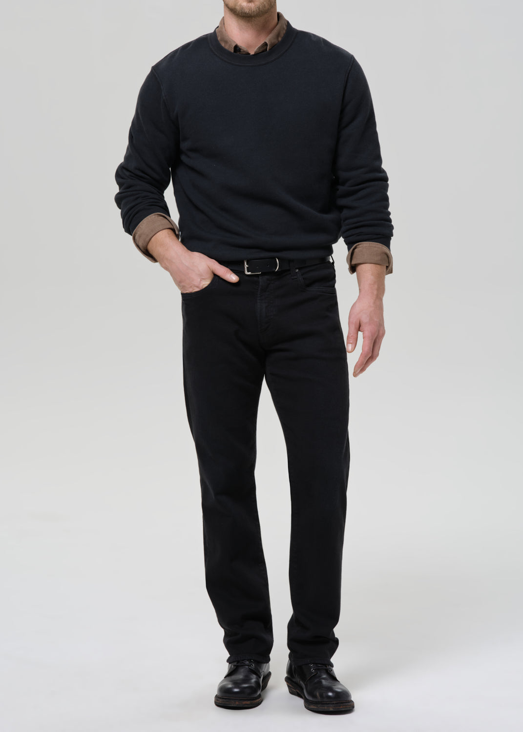 Person wearing a black crewneck sweatshirt with black straight leg jeans on a grey background.