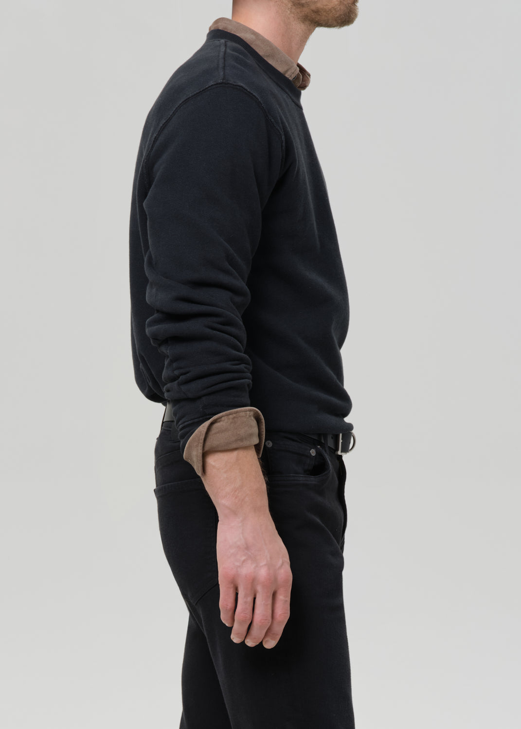 Person wearing a black crewneck sweatshirt with black straight leg jeans viewed from the side on a grey background.