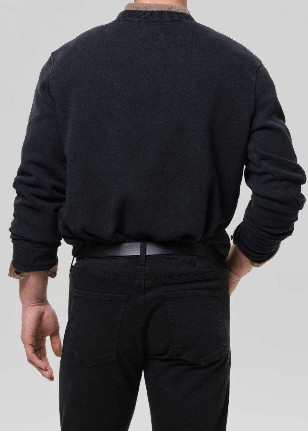 Person wearing a black crewneck sweatshirt with black straight leg jeans viewed from the back on a grey background.