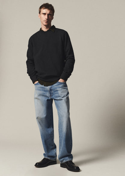 Man wearing a washed black crewneck sweatshirt layered over a dark shirt with light blue jeans on a grey background.