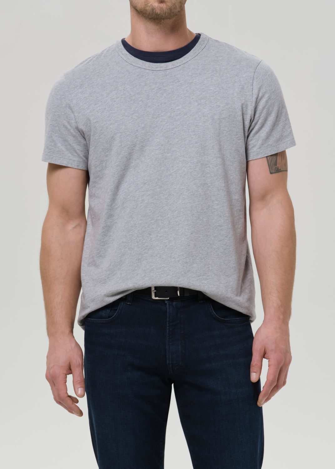 A man wearing a light grey short-sleeve crewneck t-shirt with a dark navy collar, paired with dark blue jeans, shown from the waist up against a grey background.