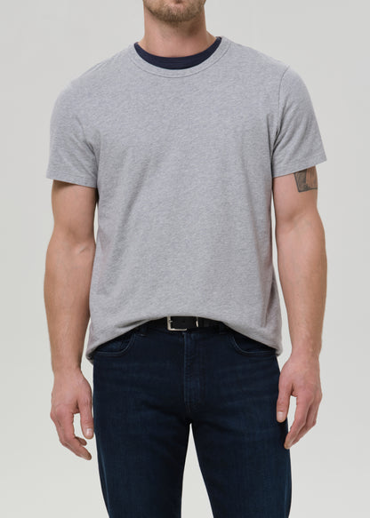 A man wearing a light grey short-sleeve crewneck t-shirt with a dark navy collar, paired with dark blue jeans, shown from the waist up against a grey background.