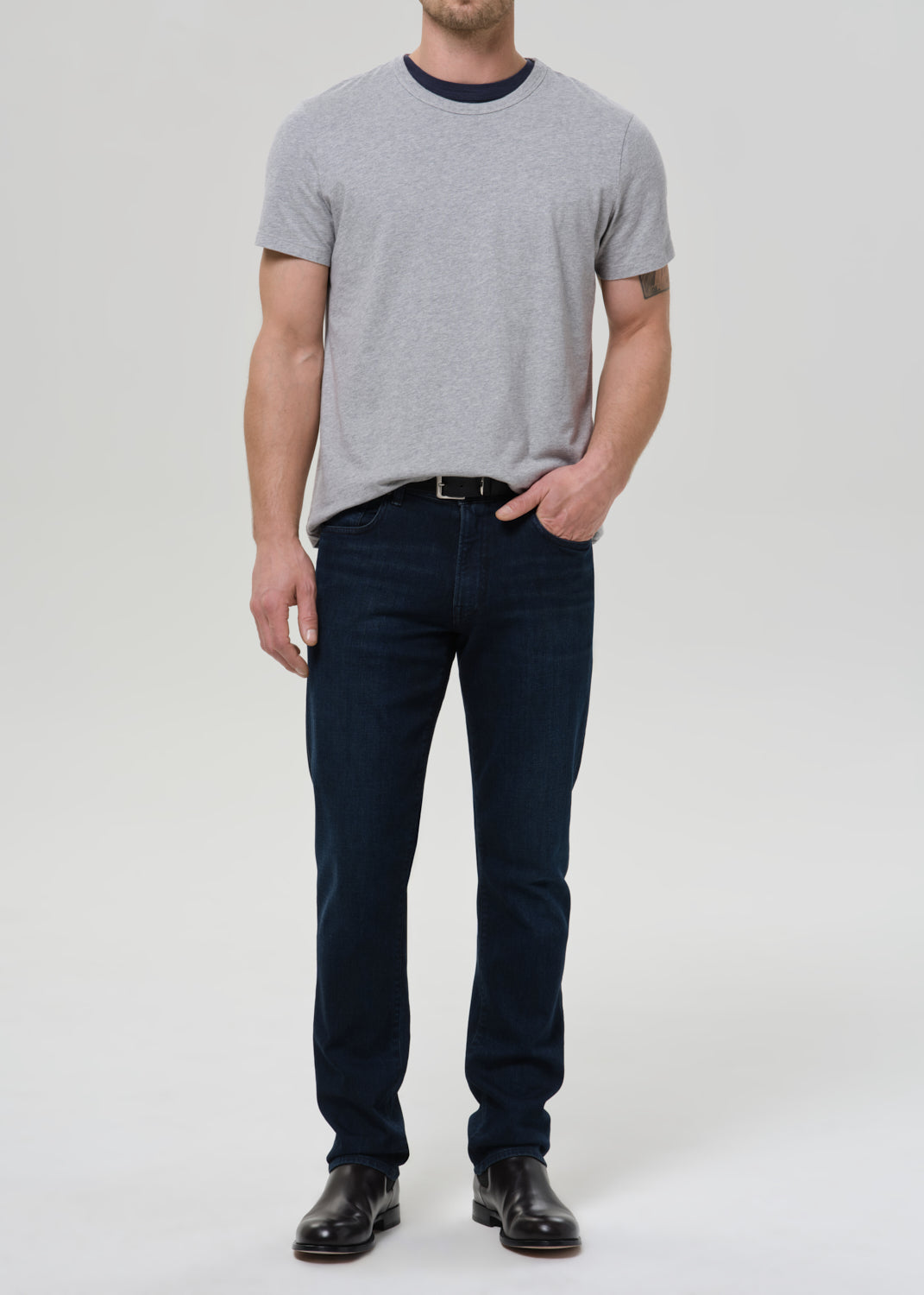 A man modeling a light grey crewneck t-shirt with a dark navy collar, paired with dark blue jeans and black leather shoes, standing against a light grey background.