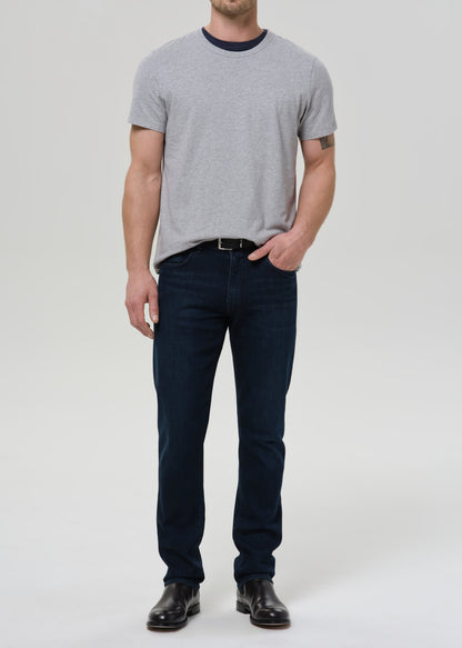 A man modeling a light grey crewneck t-shirt with a dark navy collar, paired with dark blue jeans and black leather shoes, standing against a light grey background.