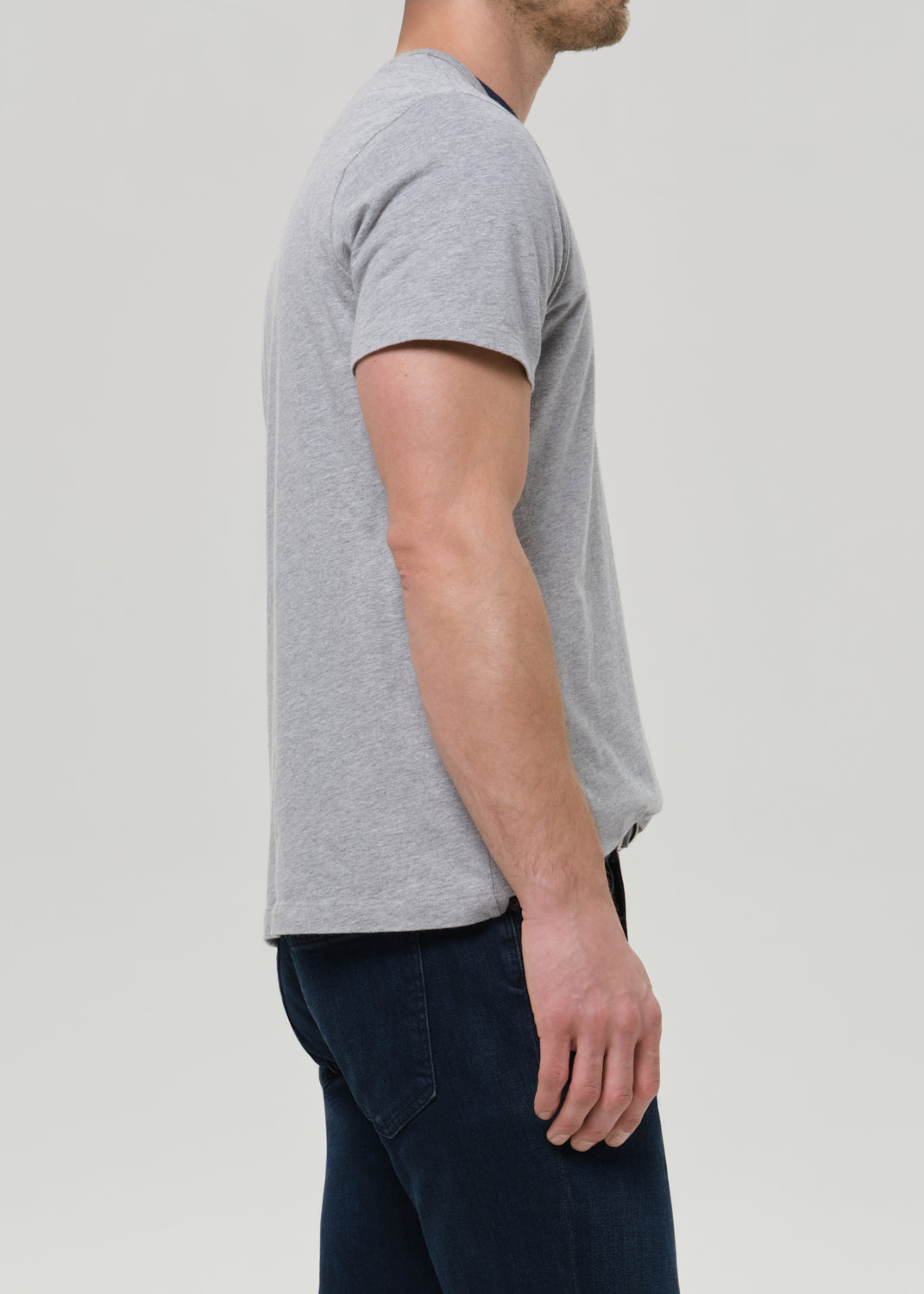 A man shown in side profile wearing a light grey short-sleeve crewneck t-shirt and dark blue jeans, photographed against a solid light grey background.