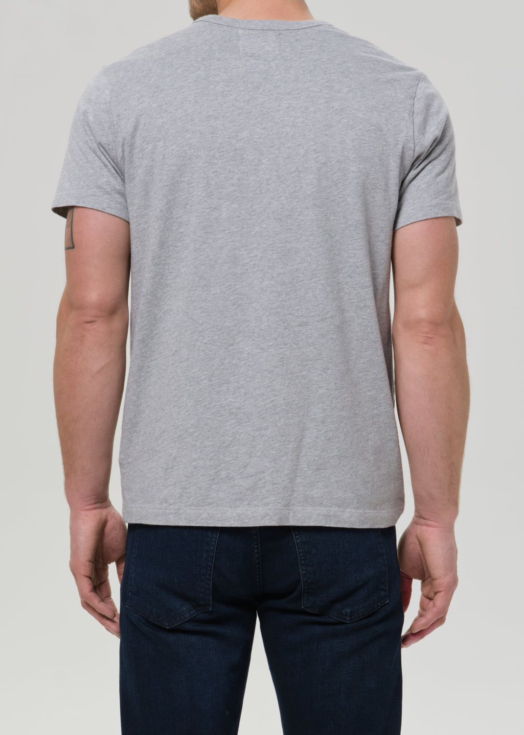 A man viewed from the back wearing a light grey heather crewneck t-shirt and dark blue jeans, set against a grey background.